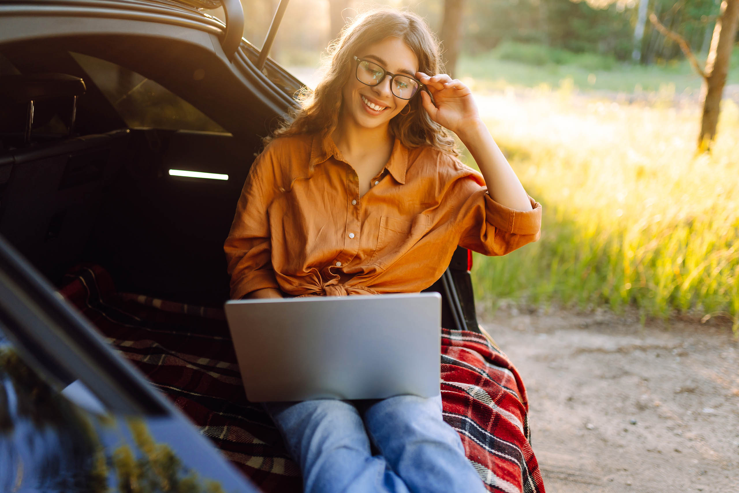 Young woman sitting in the trunk of a car with a laptop on a sun